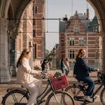 Women cycling under an Amsterdam archway with historical buildings in the background.