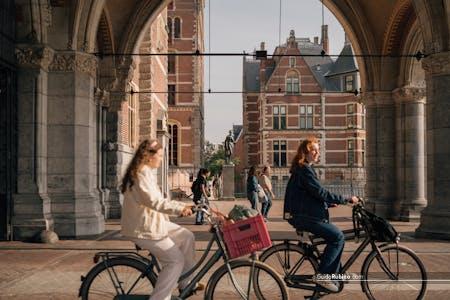 Women cycling under an Amsterdam archway with historical buildings in the background.